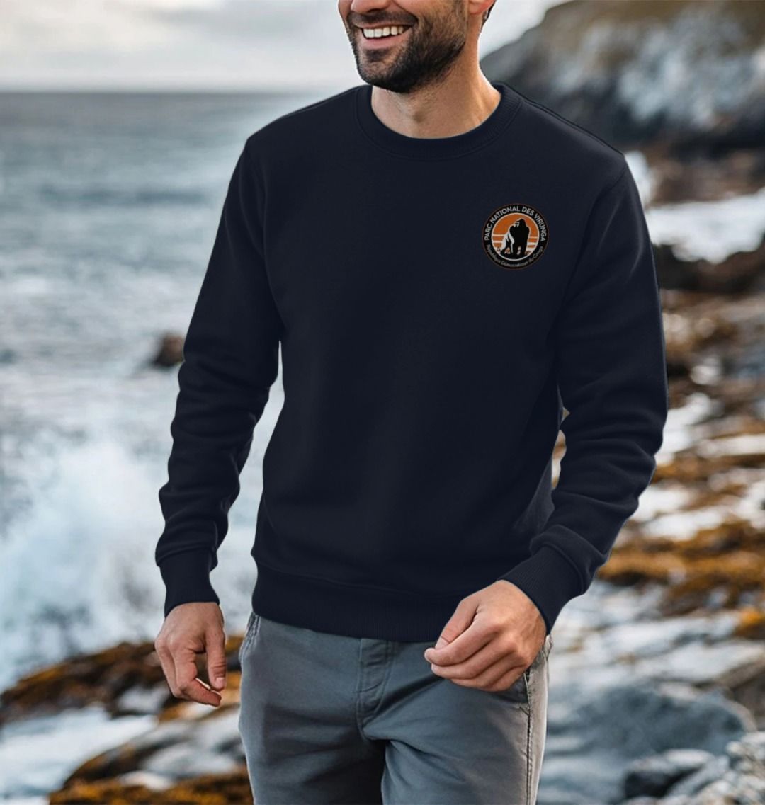 A man standing on a rocky beach wearing a black Virunga Pocket Logo Men's Sweater