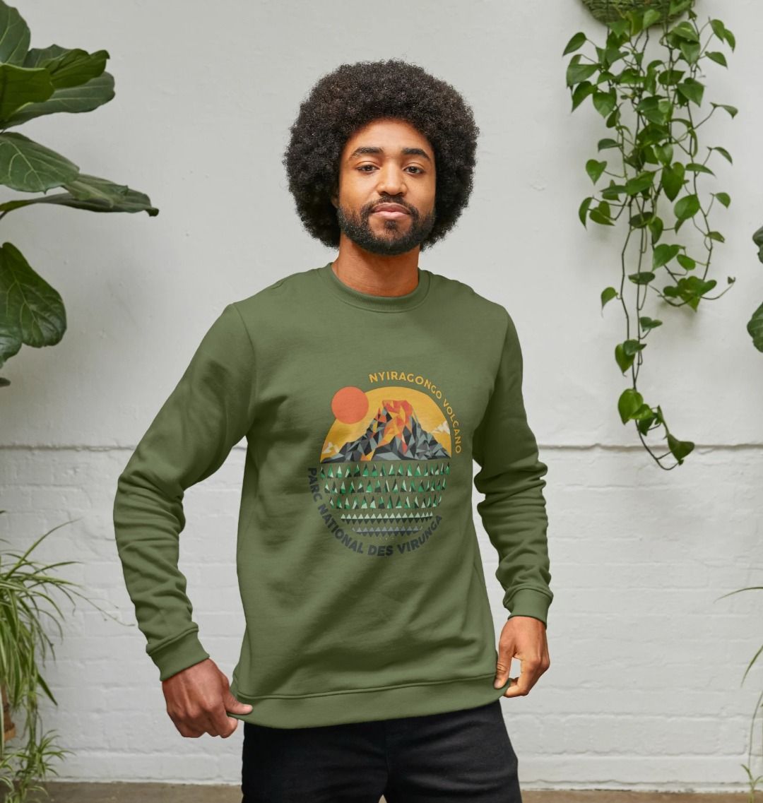 A man wearing a khaki Nyiragongo Volcano Men's Sweater, standing against a light gray wall with plants.