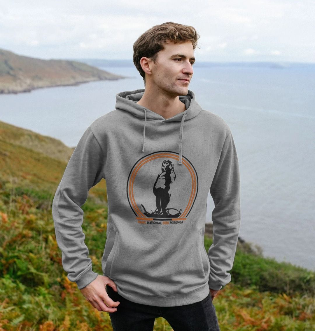 A man wearing a light heather Take A Stand Men's Hoodie standing on a coastal landscape.