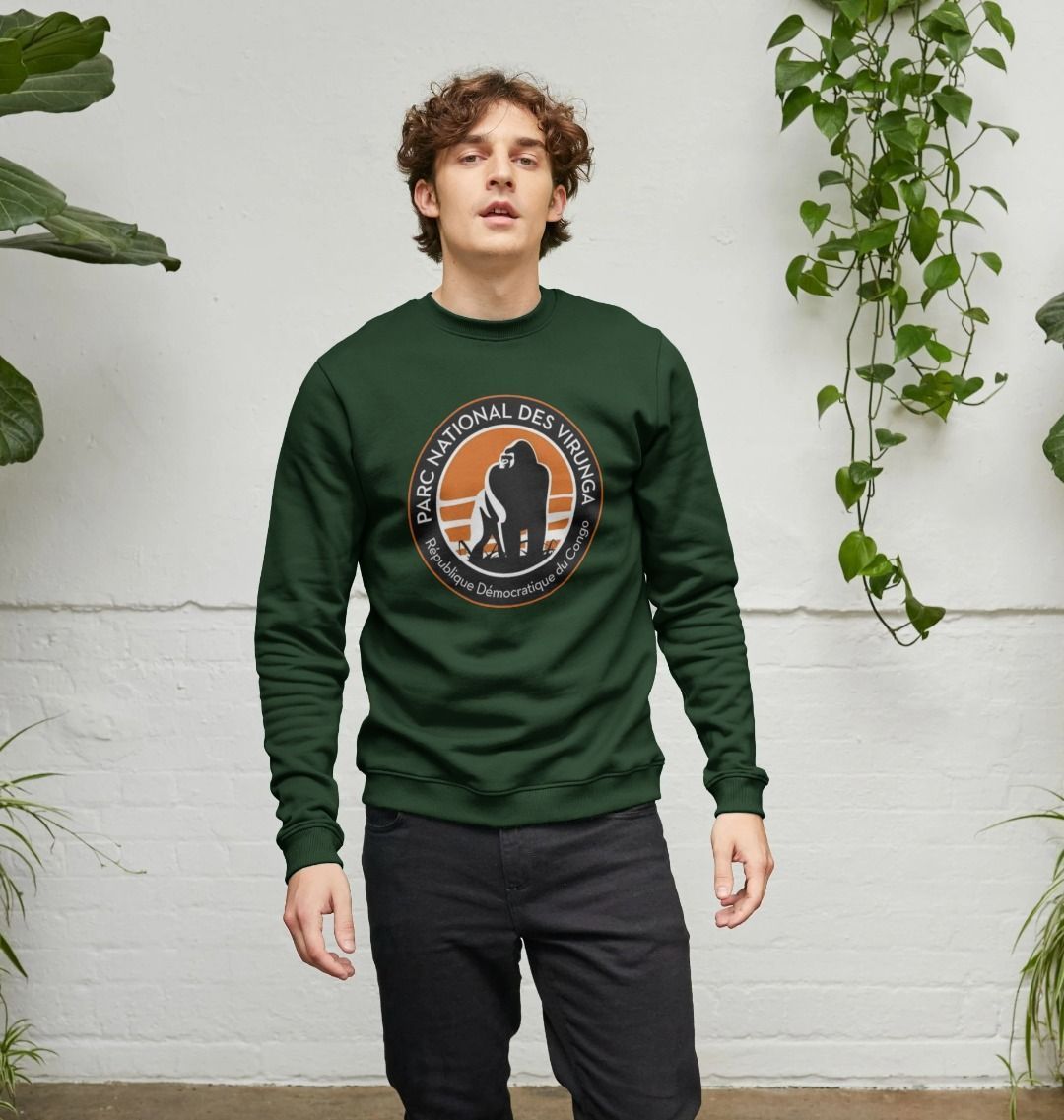 A man wearing an evergreen Virunga Logo Men's Sweater against a white wall with plants.