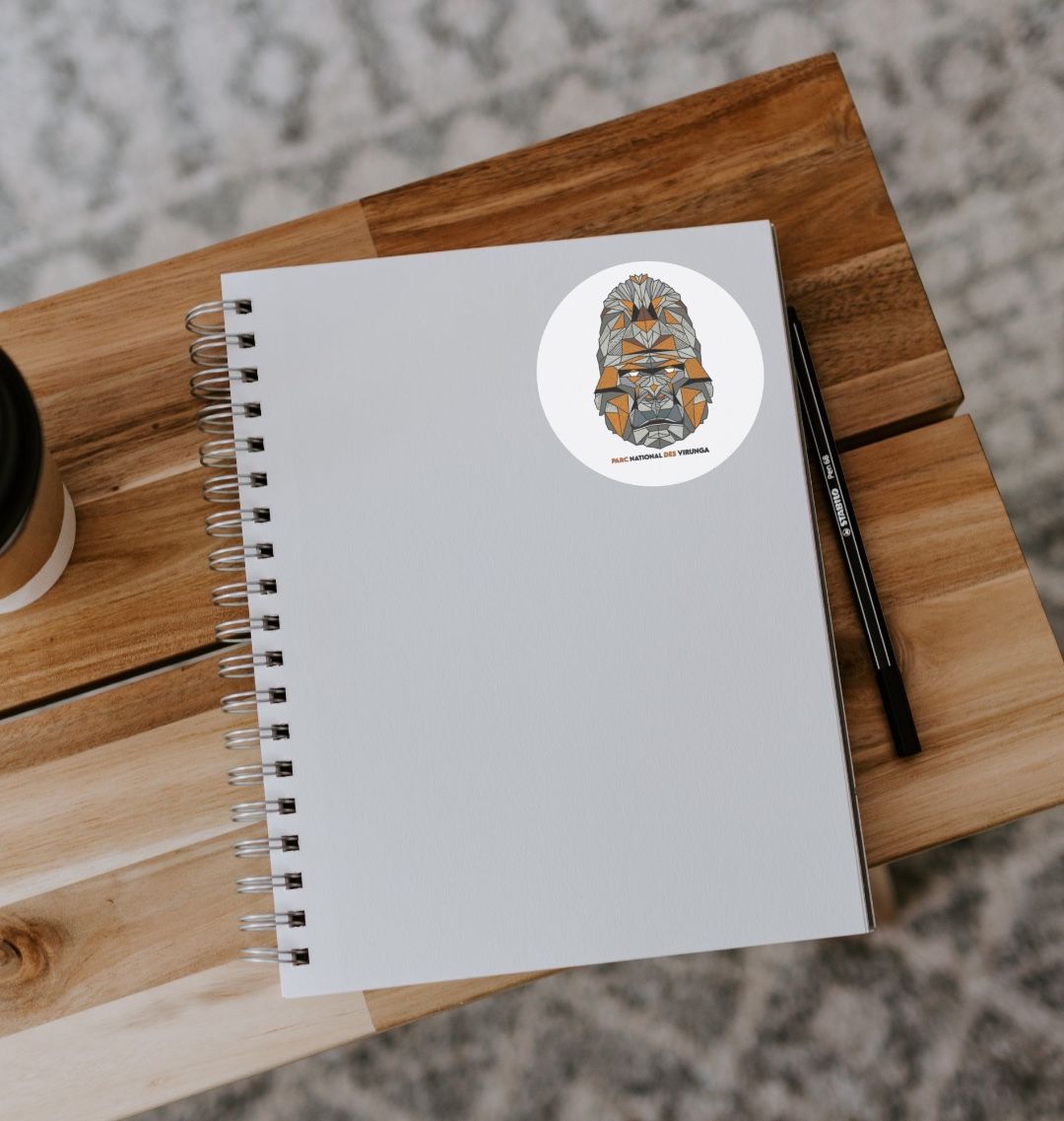 A white spiral notebook with a A White Gorilla Face Sticker on its cover, displayed on a wooden table next to a pen and a cup.