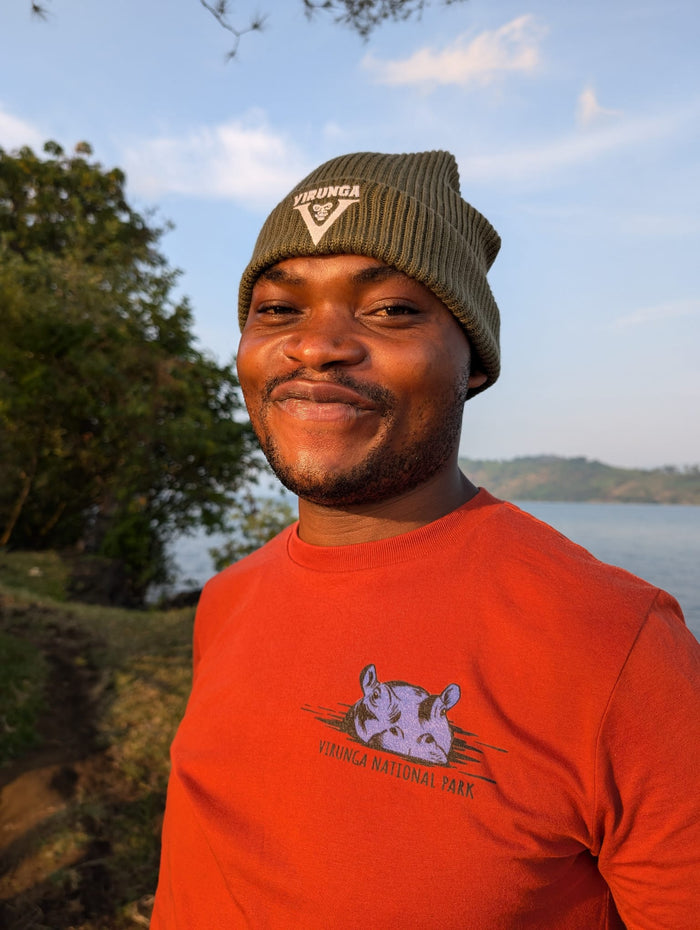 A person wearing a green Virunga beanie hat and a rust Virunga Hippo T-shirt, standing near a body of water in the Virunga National Park (Tchegera Island).