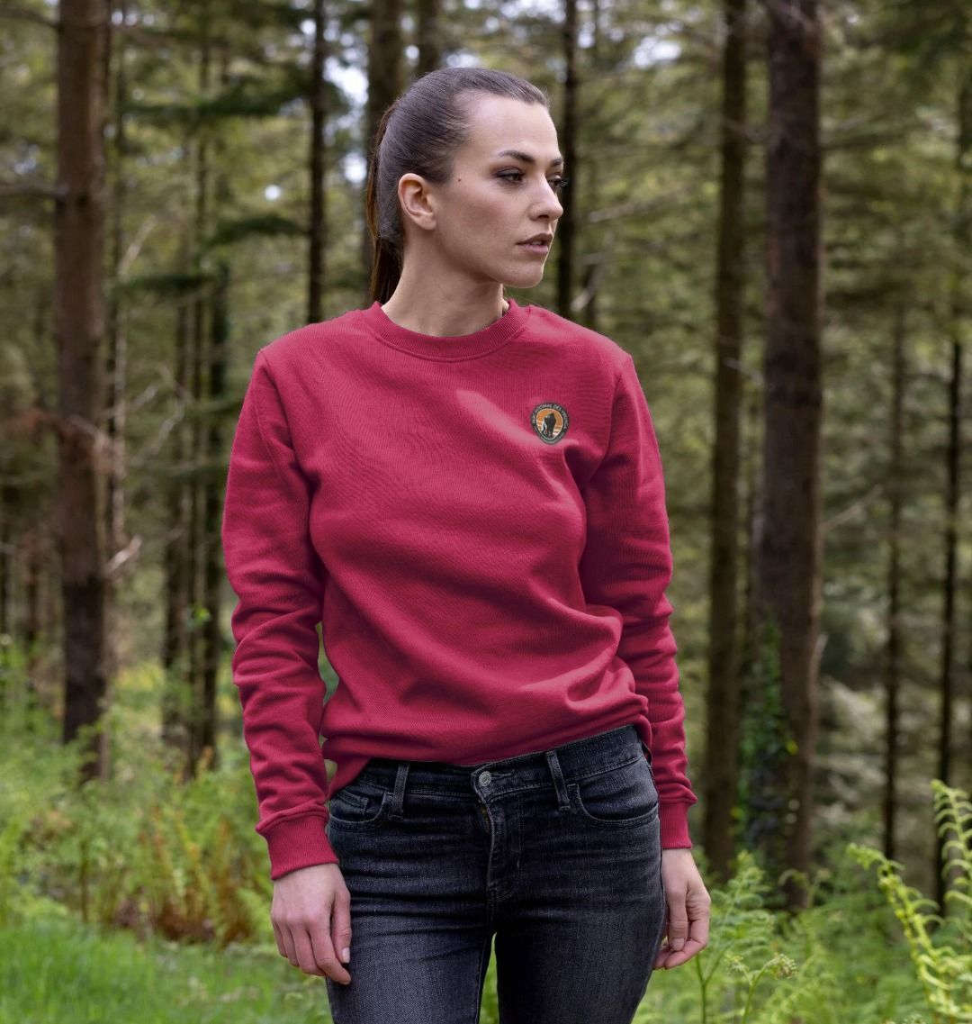 A woman standing in a forest wearing a cherry Virunga Pocket Logo Women's Sweater, paired with black pants.