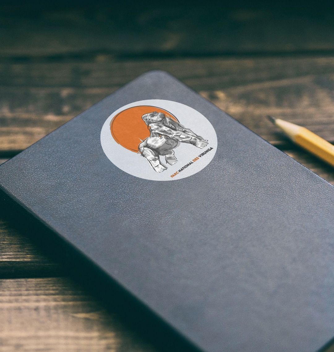 A gray notebook with a Ndakasi Sticker on the cover, placed on a wooden surface next to a pencil.