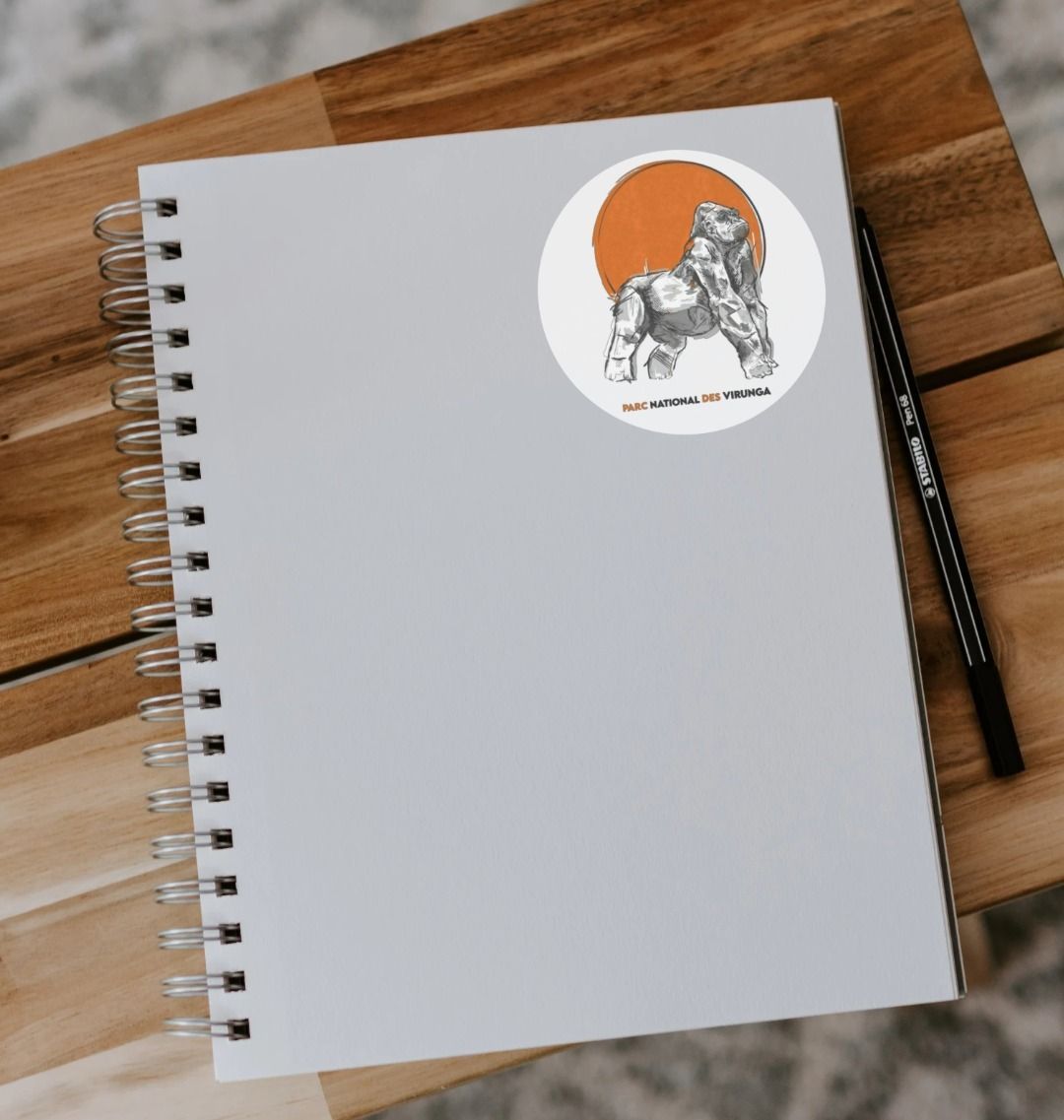 A grey notebook with a Ndakasi Sticker on the cover, placed on a wooden surface next to a pencil.