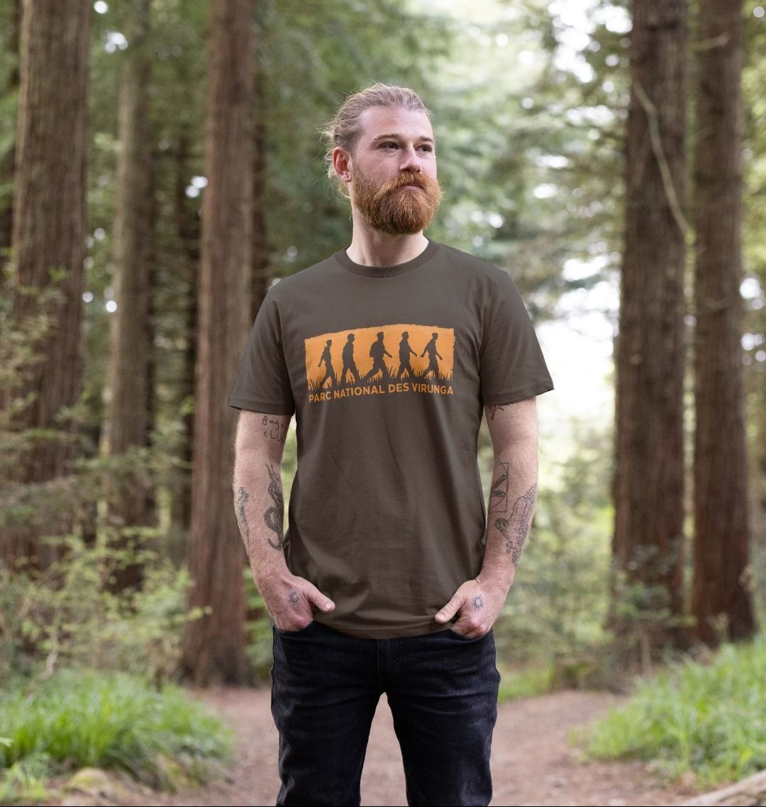 A man standing in a forest wearing a Chocolate Virunga Rangers Men's T-shirt with a mountainous backdrop.