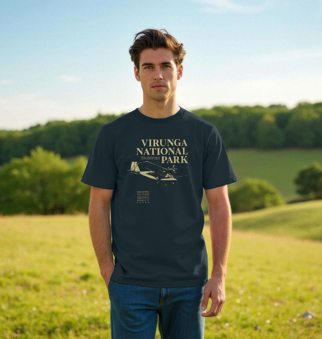 A man wearing a denim blue Virunga Pilot Men's T-shirt, standing in a grassy field with trees in the background.