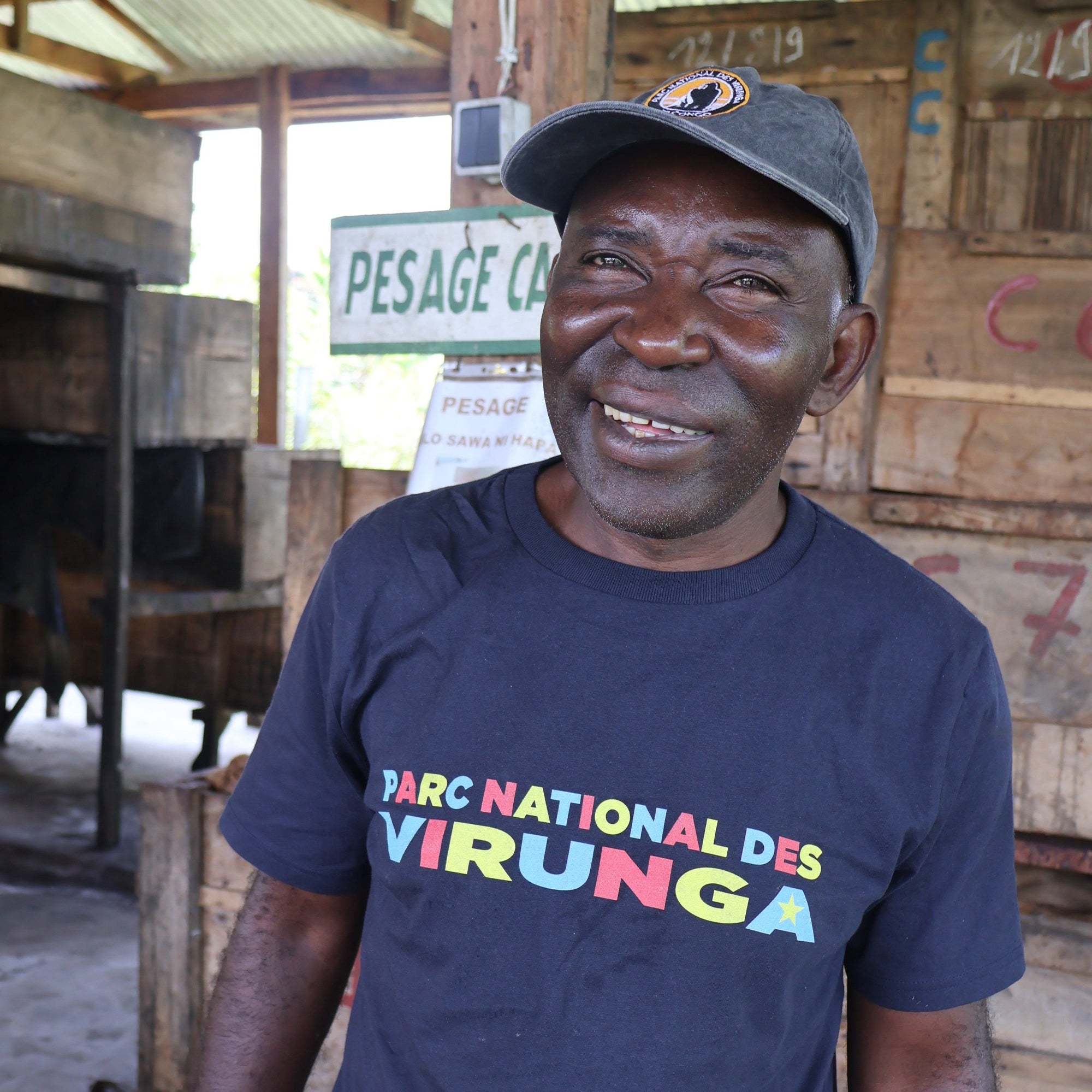 A man wearing a navy blue Congo Heritage Men's T-shirtt, standing in front of a wooden structure with a sign that reads 'PESAGE'.