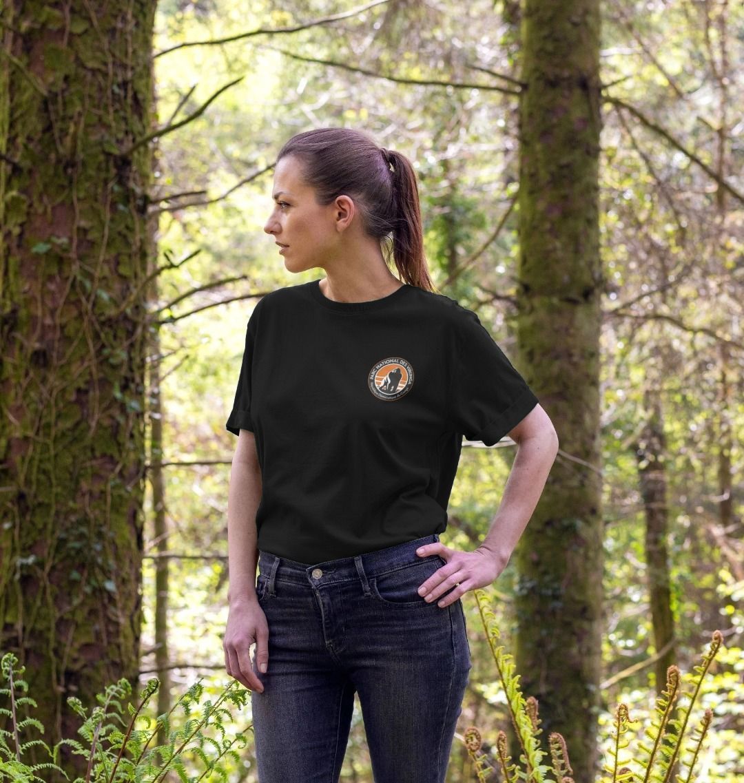 A woman standing in a forest setting, wearing a black Women's Okapi T-shirt.
