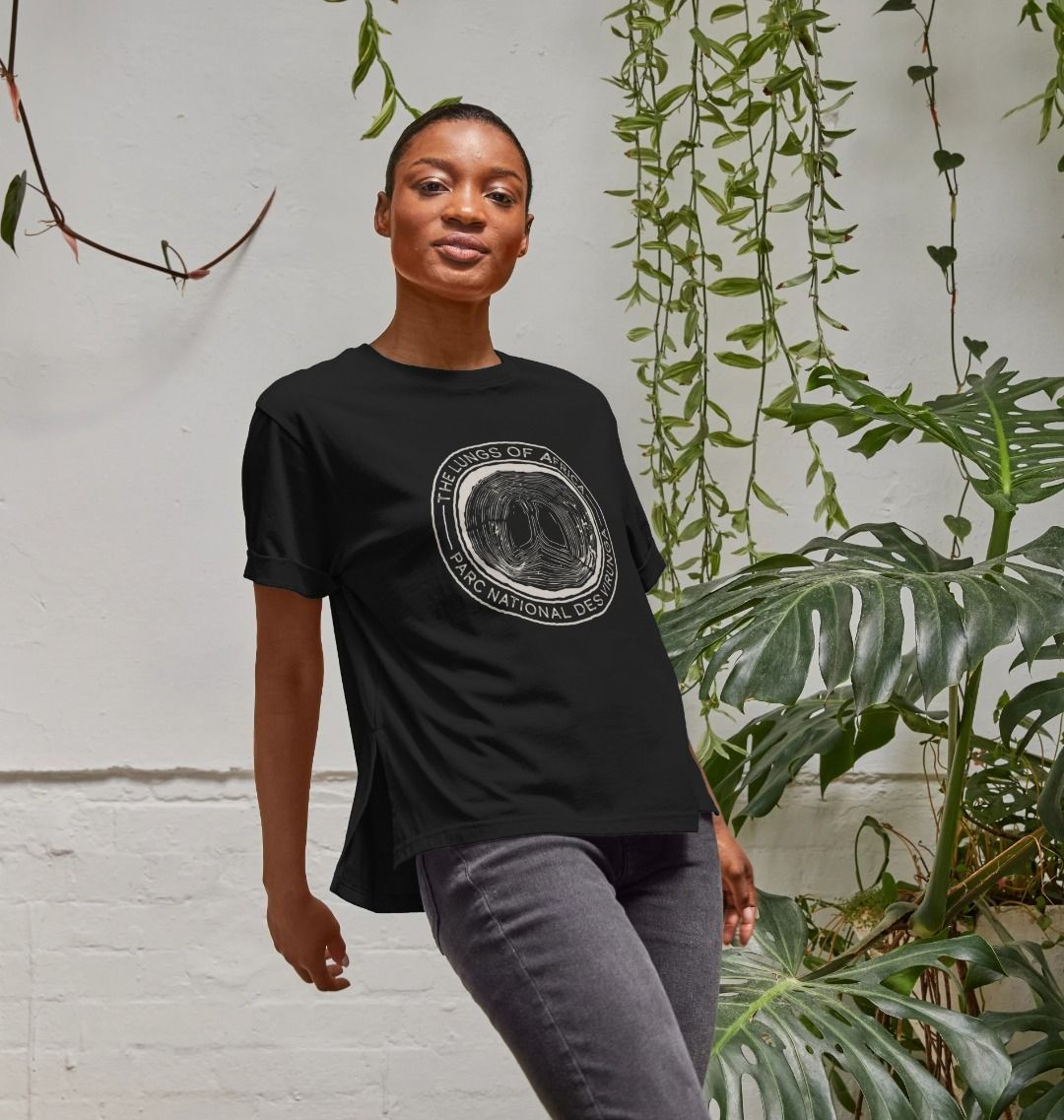 A woman standing in front of a white wall with plants, wearing a black Lungs of Africa Women's Relaxed Tee.