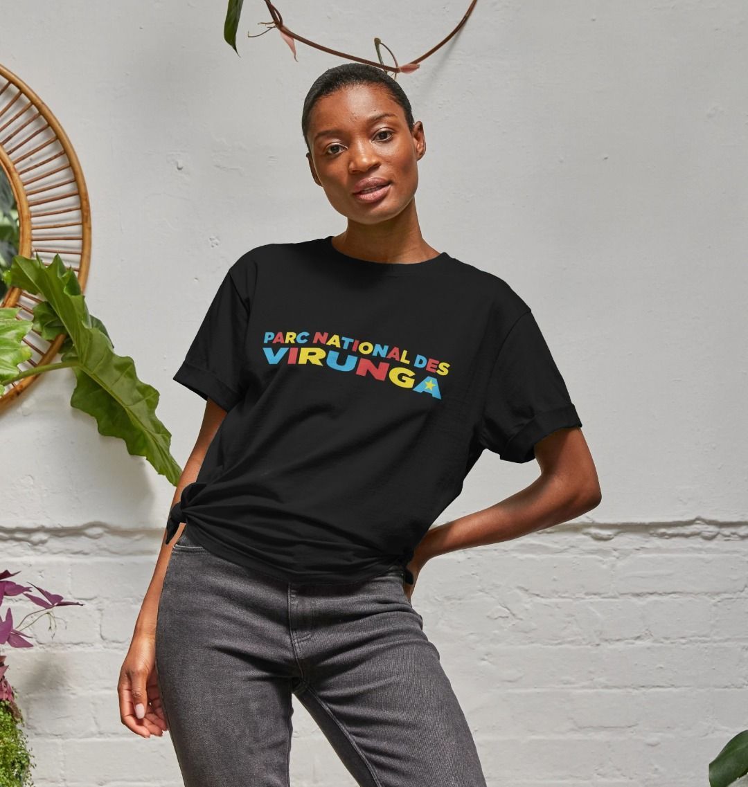 A woman wearing a black Congo Heritage Women's Relaxed Tee, standing in a room with a brick wall and plants in the background.