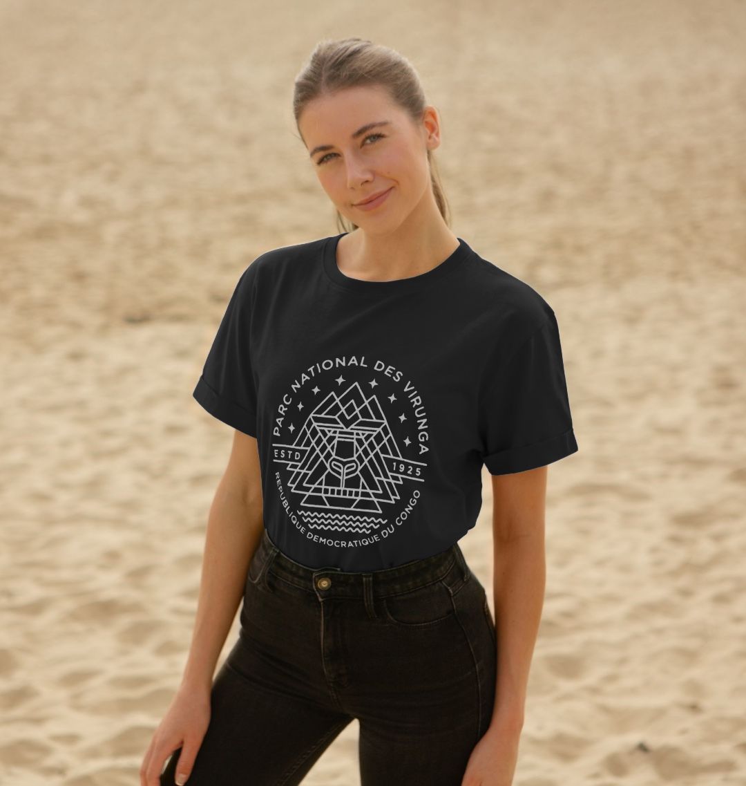 A woman standing on a sandy surface wearing a black Virunga Emblem Women's Relaxed Tee, and dark pants.
