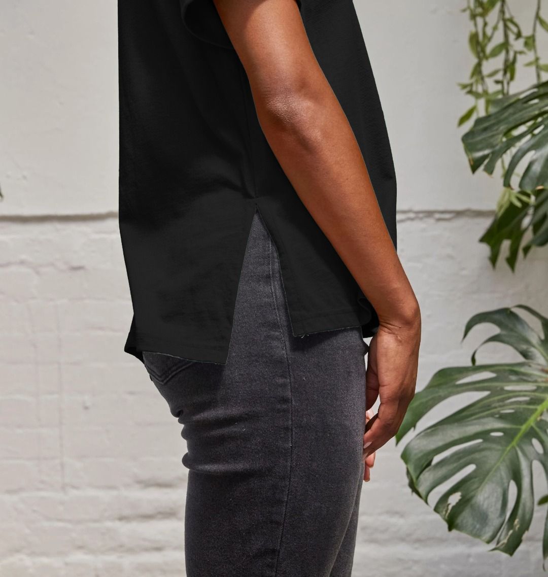 A woman standing in front of a white wall with plants, wearing a black Lungs of Africa Women's Relaxed Tee and black pant