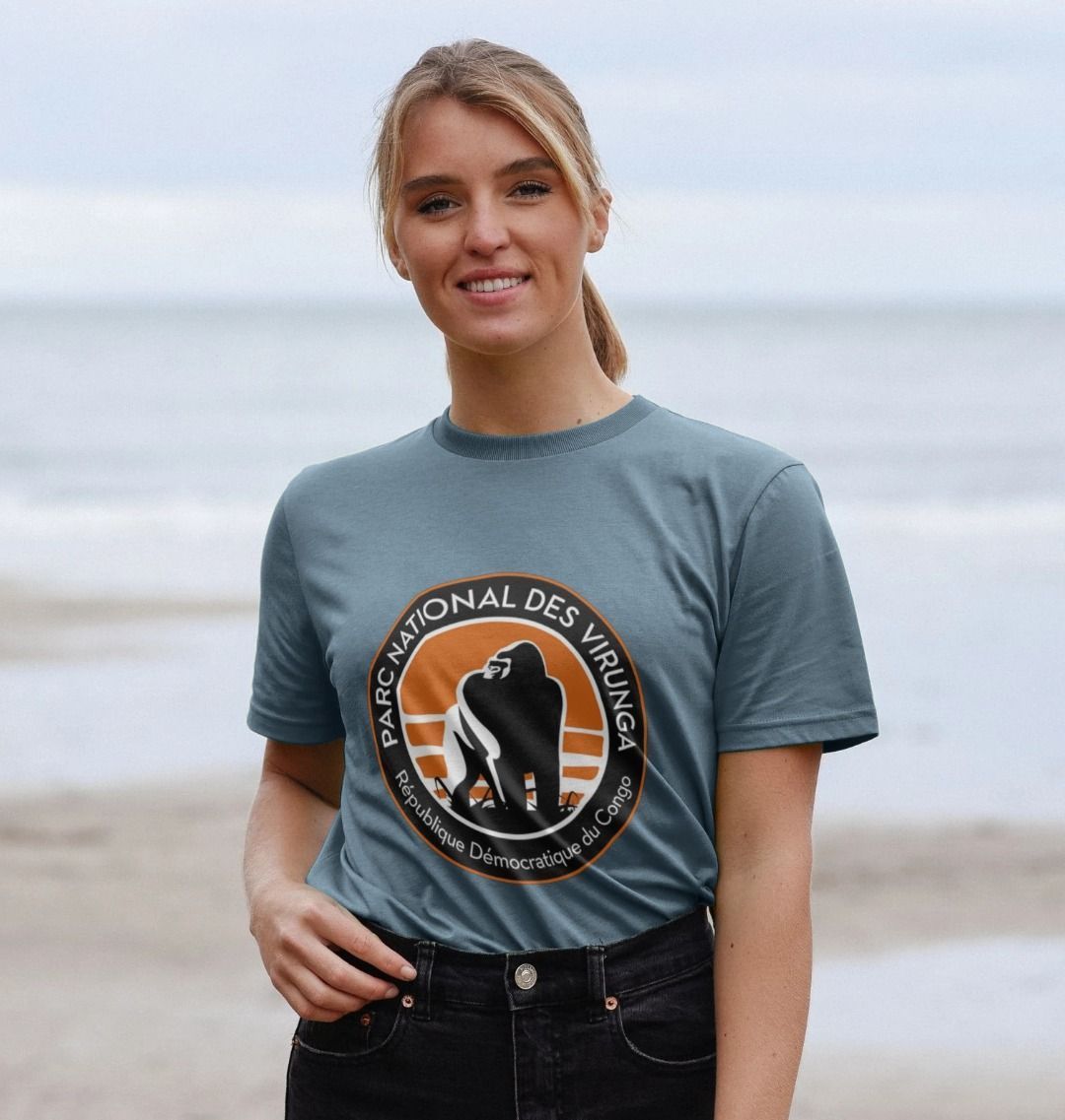 A woman wearing a blue Virunga Logo Women's T-shirt, standing near a body of water.