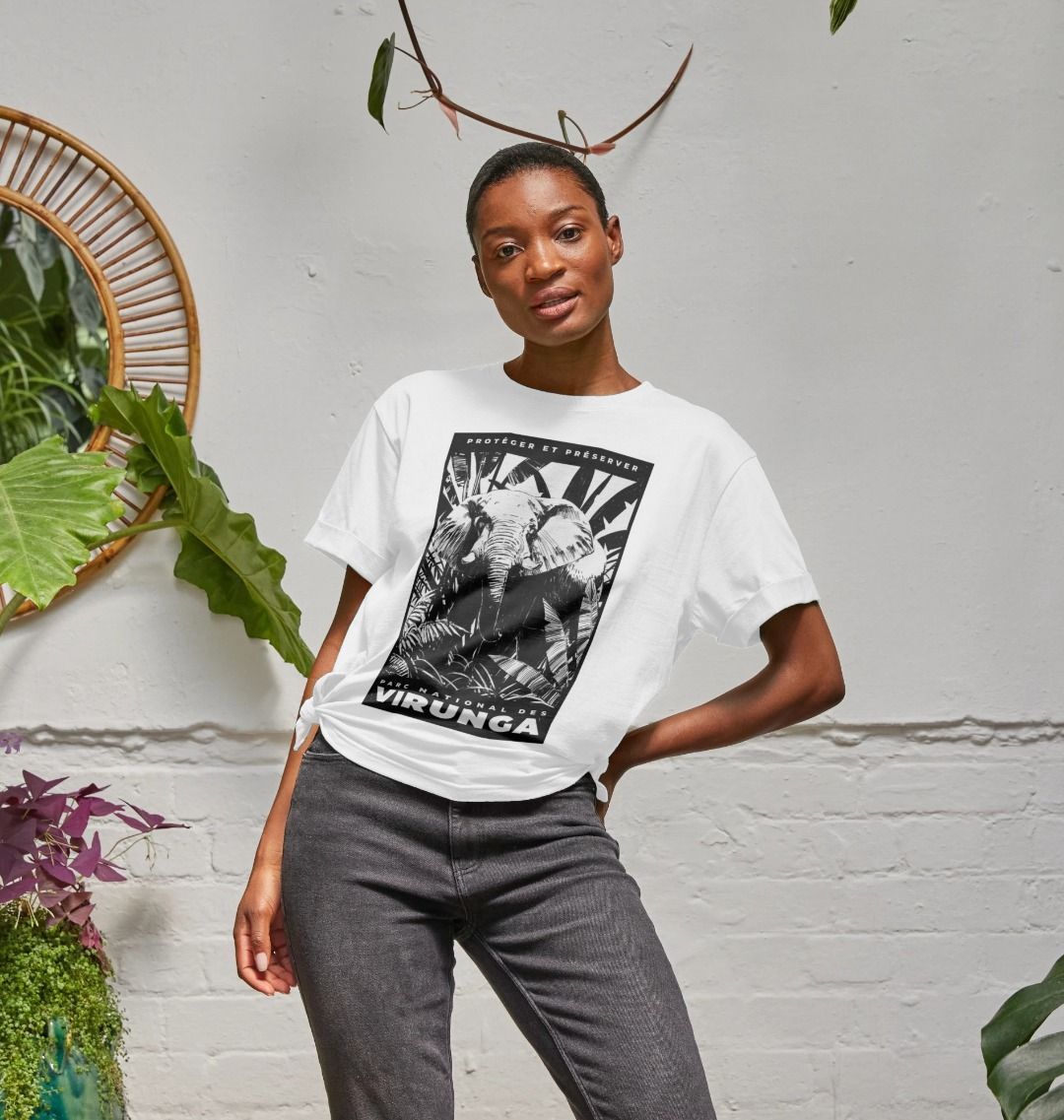 A woman wearing a white African Elephant Black Print Women's Relaxed Tee, standing in front of a white wall with plants.