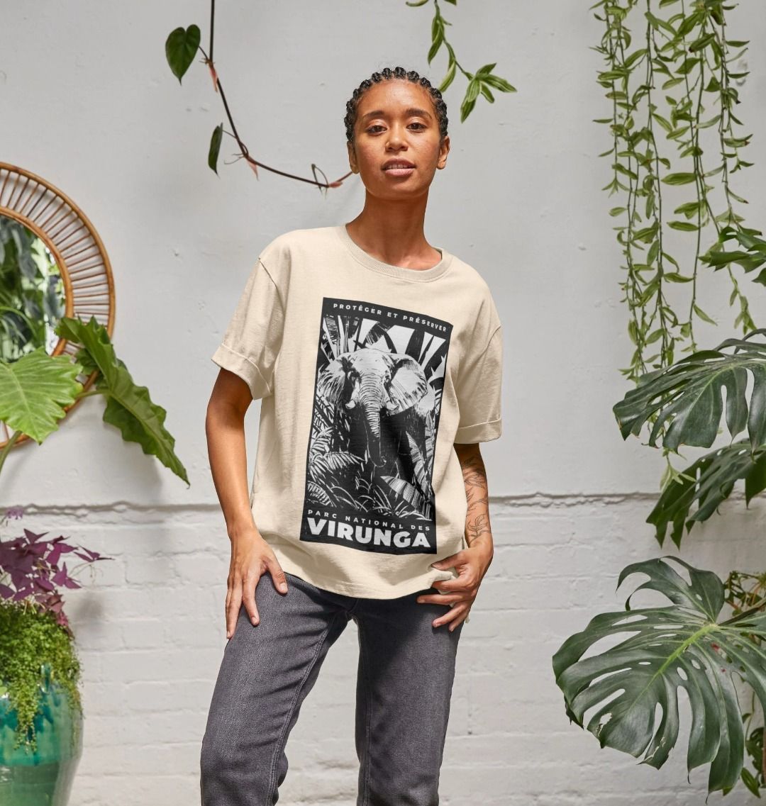 A woman wearing an oat African Elephant Black Print Women's Relaxed Tee, standing in front of a white wall with plants.