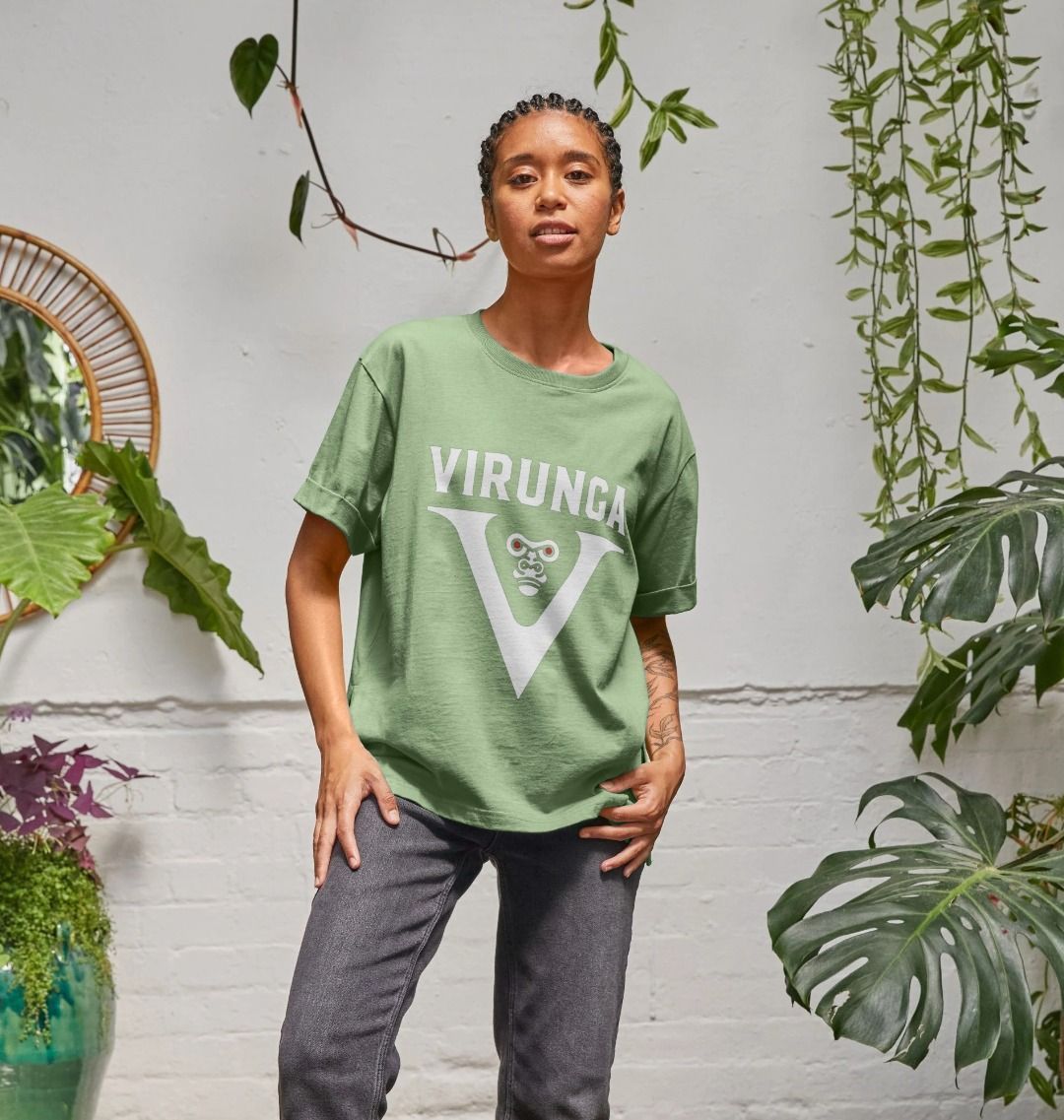 A woman standing in a room with plants, wearing a sage V for Virunga Women's Relaxed Tee and a black pant