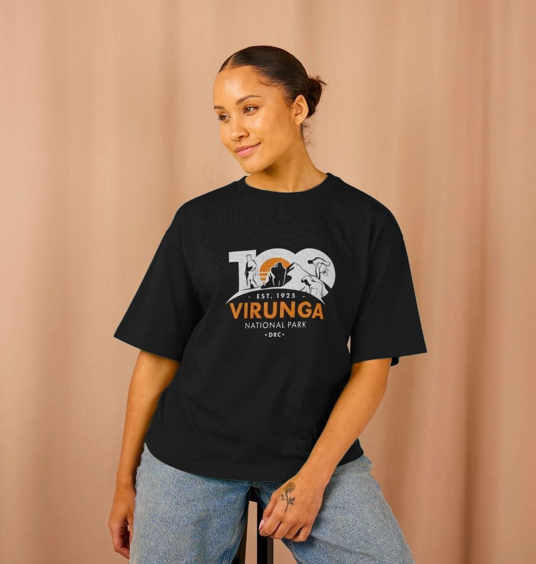 A woman wearing a black Virunga 100 Women's Oversize T-shirt standing against a beige background.