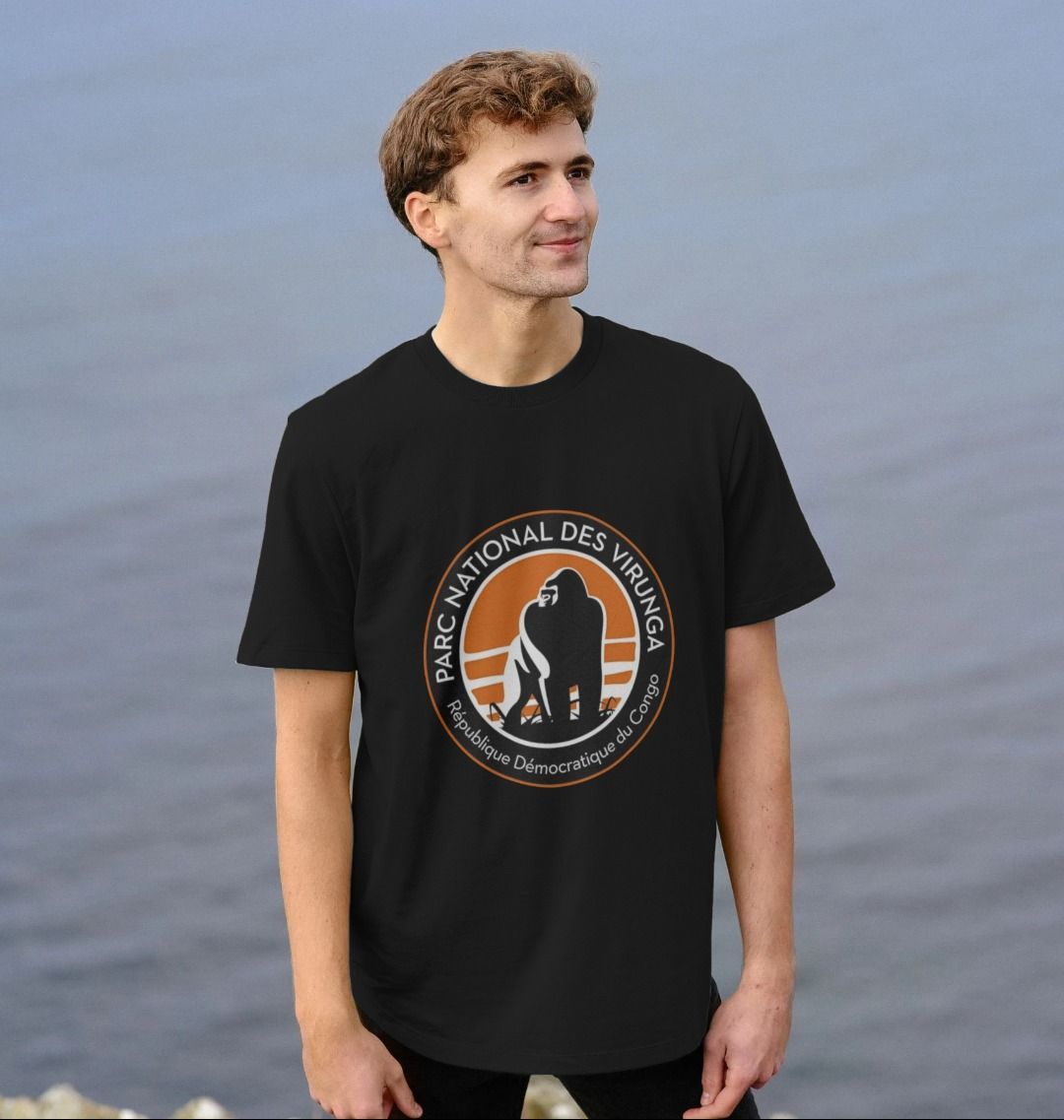 A man standing by a body of water wearing a Virunga logo black longline t-shirt with.